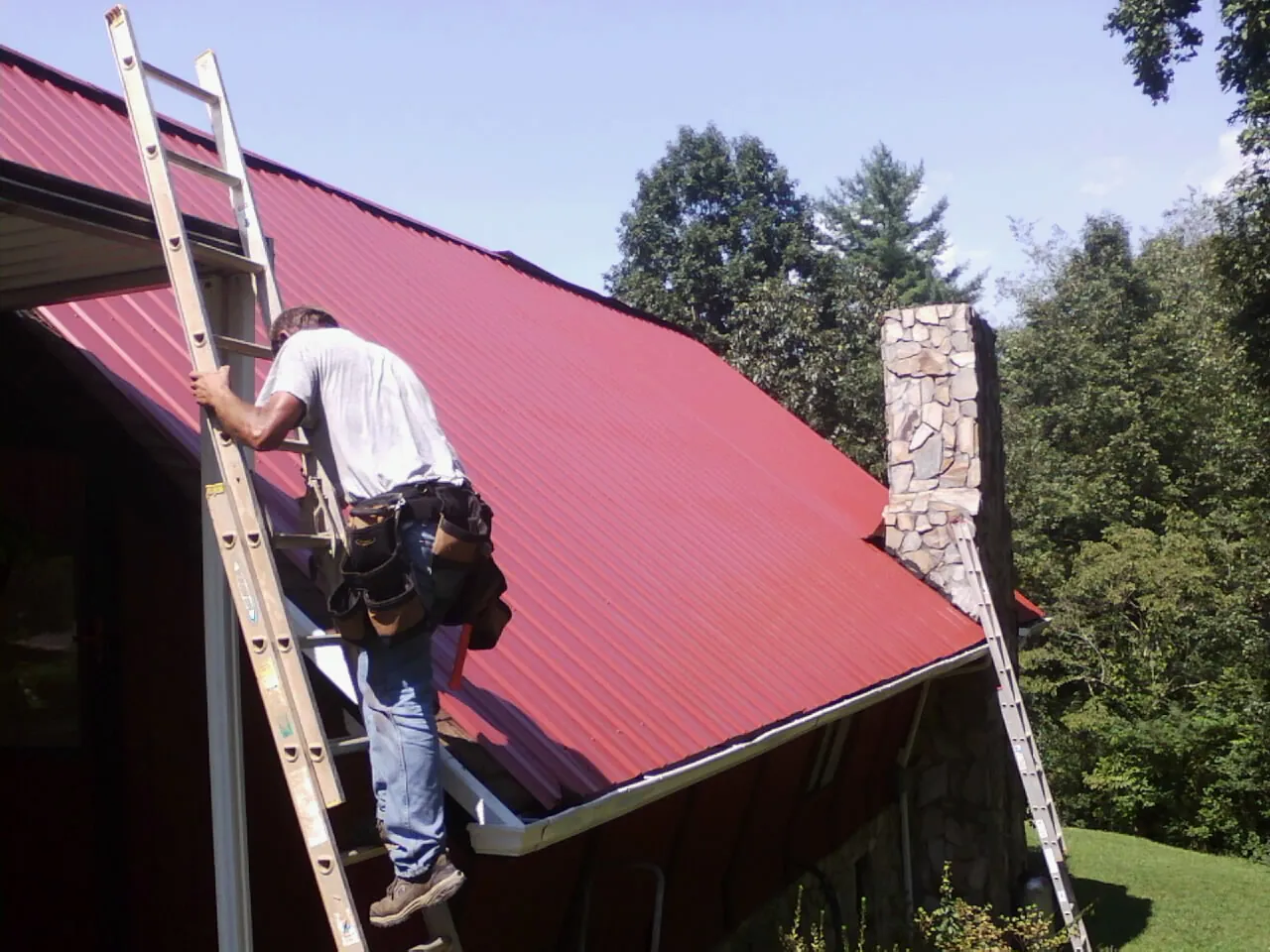 Roofer on a ladder beside a red metal roof and stone chimney — Soffit & Fascia Repair by Roofing Brothers in Ellerbe